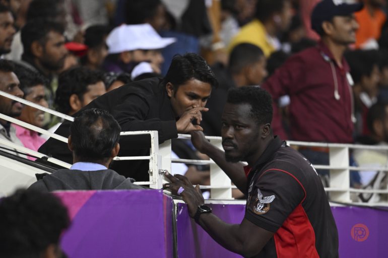 A man in a black shirt talks to a sports team staff member near a stadium crowd during a match.