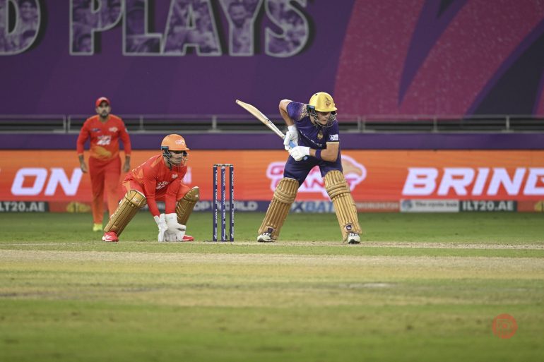 A cricket batsman plays a shot as the wicketkeeper and fielders watch during a match.