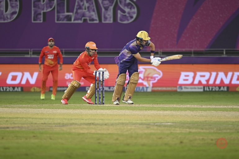 A cricket batsman in purple plays a shot as the wicketkeeper and fielders in orange look on.