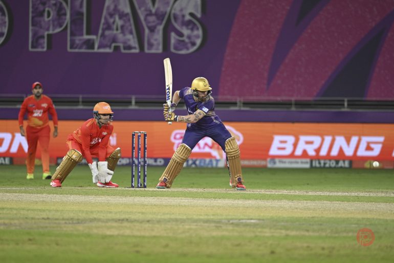 A cricket batsman in purple and gold prepares to hit the ball as the wicketkeeper and fielder watch.
