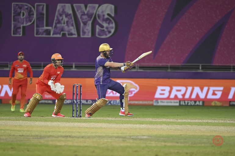 A cricket batter in purple plays a shot as the wicketkeeper and fielders in orange watch on the field.