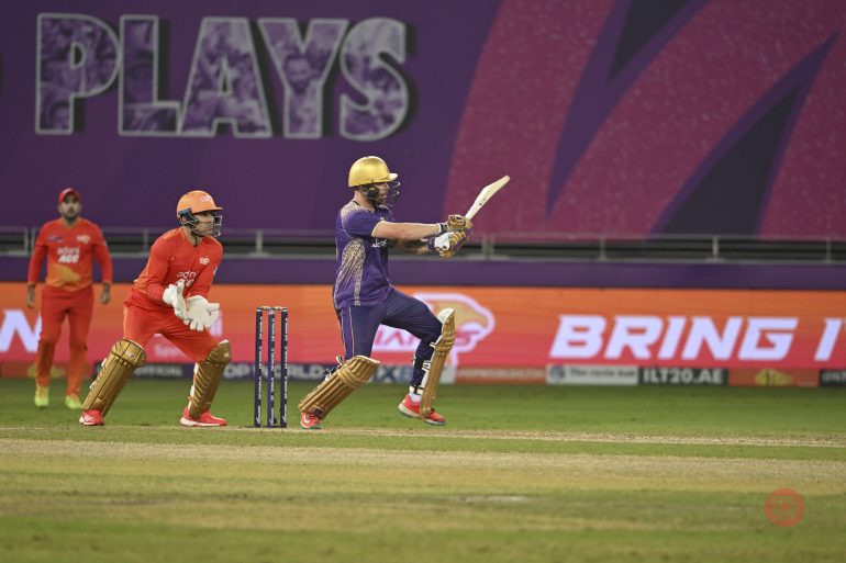 A cricket batsman in purple plays a shot as the wicketkeeper and fielder in orange look on.