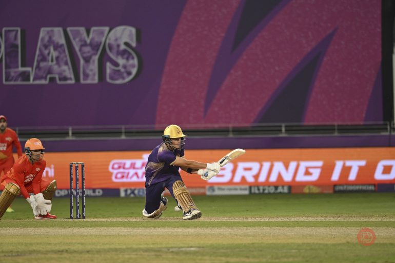 A cricket batter in purple plays a shot as the wicketkeeper in orange watches during a match.