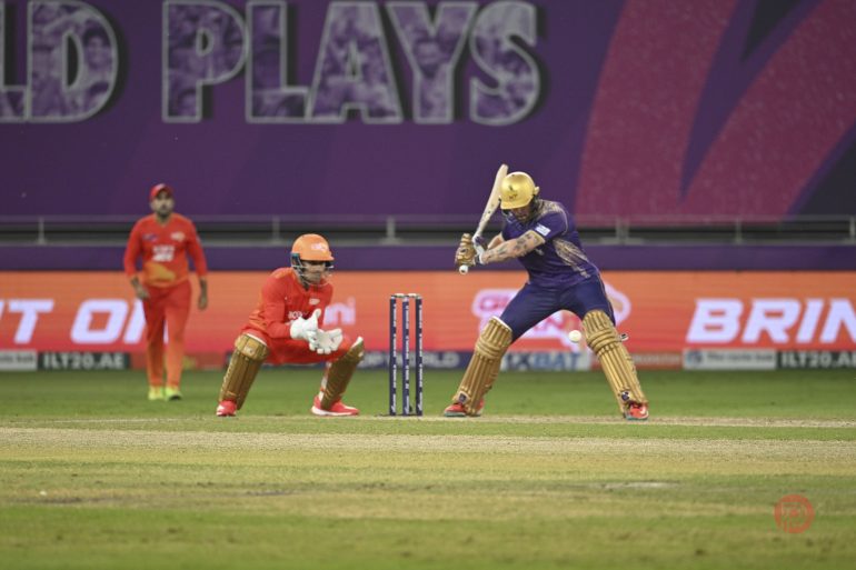 Cricketer in purple batting as wicketkeeper and fielder in orange watch during a match on a stadium field.