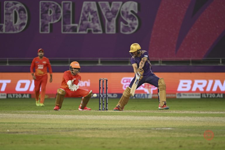 A cricket batsman plays a shot as the wicketkeeper and a fielder watch closely on a brightly lit field.