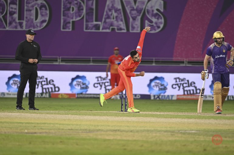 A cricket bowler in an orange uniform delivers a ball as the umpire and batsman look on.