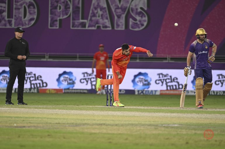 A cricket bowler in orange delivers the ball as the batsman in purple and a referee look on.