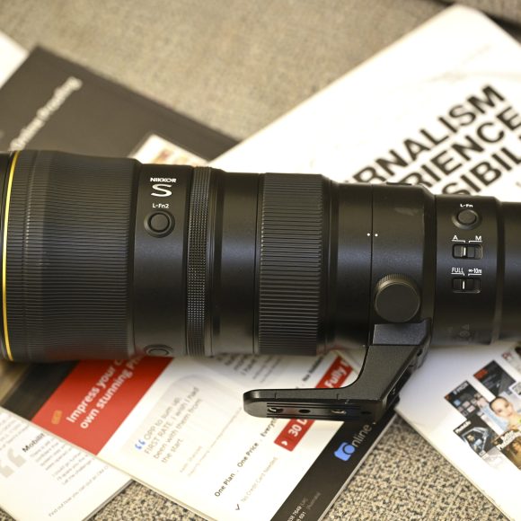A Nikon telephoto camera lens rests on magazines and papers on a desk.