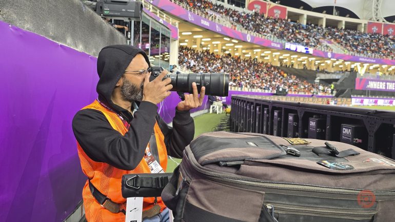 A photographer in an orange vest takes pictures at a crowded stadium event, sitting near the field.