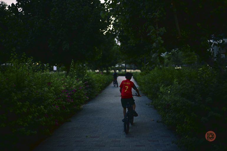 A person in a red shirt riding a bicycle down a tree-lined path, followed by another cyclist.