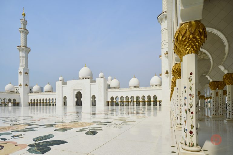 White marble mosque courtyard with floral designs, domes, and ornate golden columns under a clear blue sky.