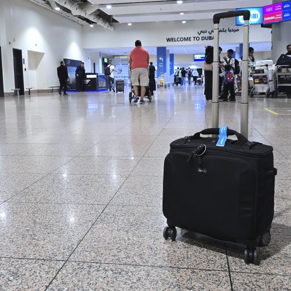 A black suitcase on wheels sits on the floor of an airport arrival area in Dubai, with people in the background.