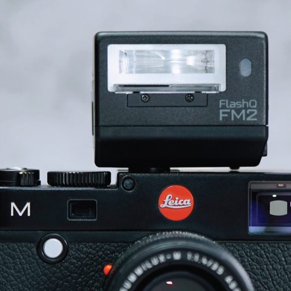 A close-up of a Leica camera with a FlashQ FM2 flash unit attached on top.