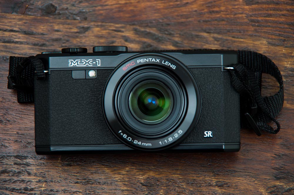A black Pentax MX-1 camera with a lens, placed on a wooden surface.