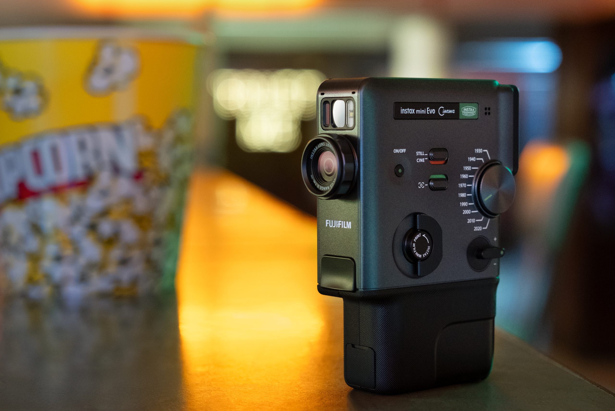 This New Fujifilm instax Camera and Printer Could Change How You Print Photos A Fujifilm Instax Mini Evo camera on a counter next to a yellow popcorn tub in a softly lit room.