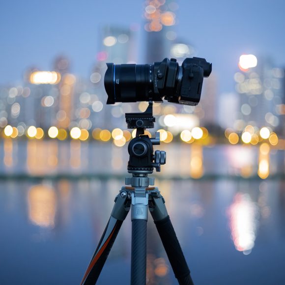 Laowa Announces The 35mm f2.8 Zero D Tilt Shift 0.5x Macro A camera on a tripod is set up by the water with a city skyline blurred in the background at dusk.