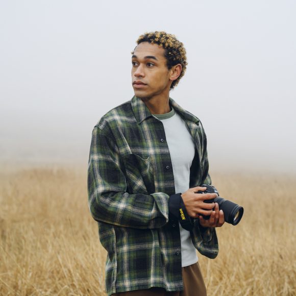 A person in a green plaid shirt holds a camera, standing in a foggy field of tall dry grass.