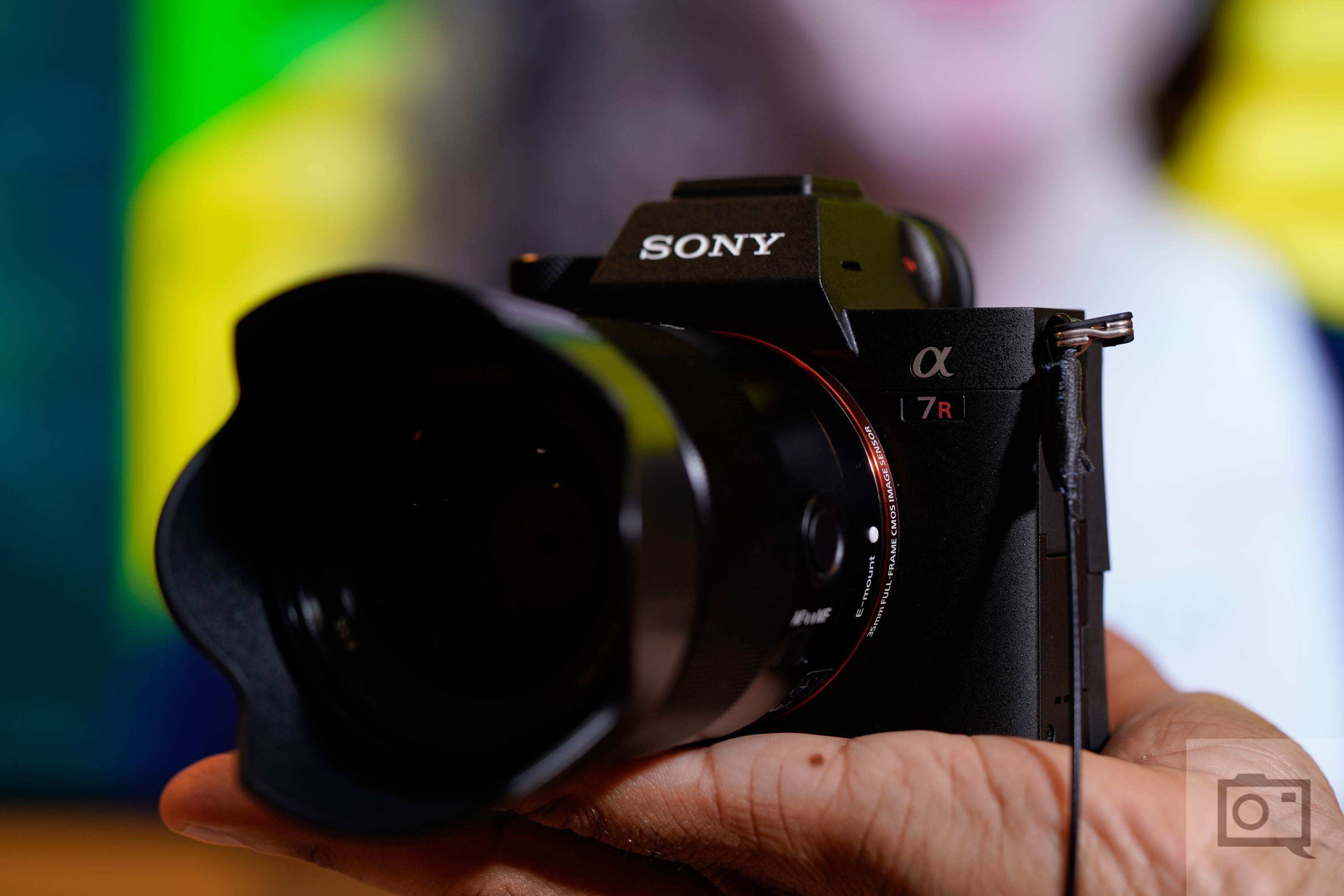 A hand holds a Sony α7R camera with a large lens, blurred background behind.
