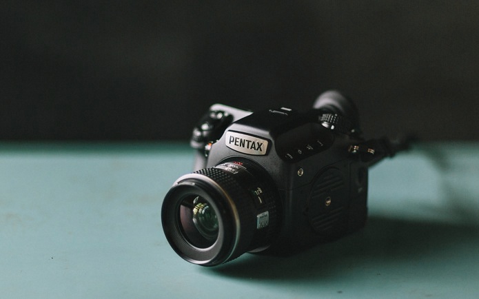 A Pentax camera with a wide lens sits on a teal surface against a dark, blurred background.