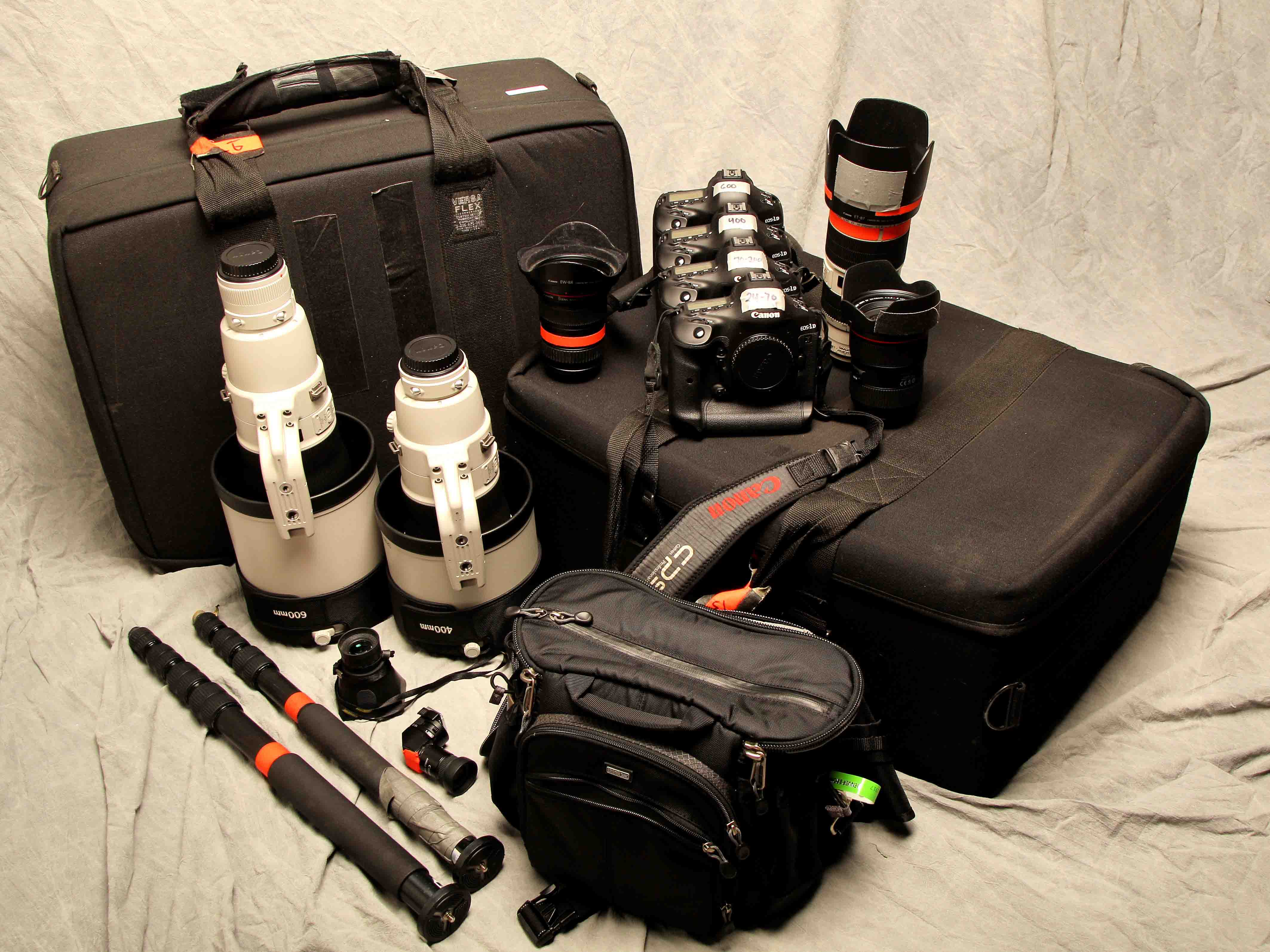 A collection of cameras, lenses, camera bags, and tripods arranged on a light fabric background.