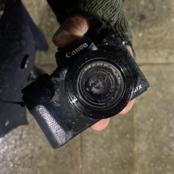 Close-up of a persons hand holding a wet Canon camera on a rainy day, with droplets on the lens and a knit glove.