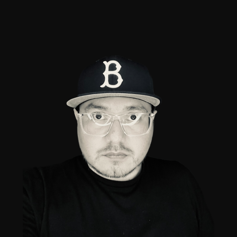 A person wearing a cap with a B logo and clear glasses, set against a black background.