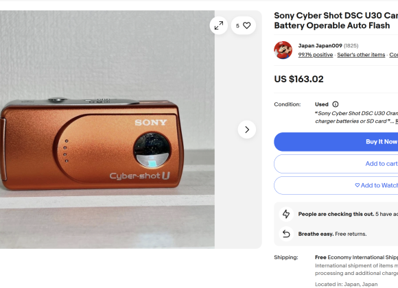 Orange Sony Cyber-shot DSC-U30 digital camera displayed on a white surface next to a wall.